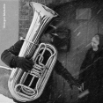 Schwarz-Weiß-Foto eines Mannes, der eine Tuba auf dem Arm trägt, während sich eine Frau nähert. Sie befinden sich mitten im Winter auf der Straße.