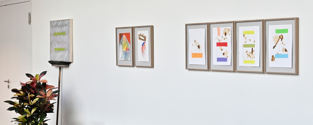 Mehrere abstrakte Gemälde an einer weißen Wand. Auf der linken Seite befindet sich eine künstlerische Installation, die aus einem umgedrehten Besen besteht, der ein abstraktes Gemälde hält.