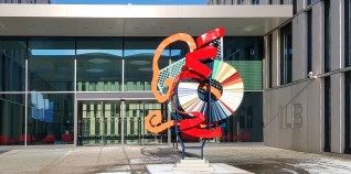 Eine aus abstrakten, farbigen Figuren hergestellte Skulptur. Die Skulptur enthällt auch einen Farbspektrum-Kreis aus Metall. Sie liegt vor dem Eingang des ILB-Gebäudes