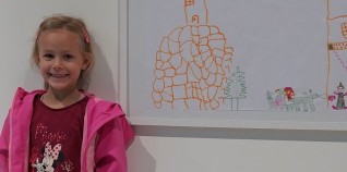 Ein kleines Mädchen lächelt. Neben ihr steht eine Zeichnung, die sie angefertigt hat.