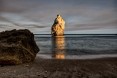 Ein Strand, dessen Meer sich in der Ferne auflöst. Im Vordergrund befindet sich ein Felsen. Im Hintergrund befindet sich ein zweiter Felsen, auf dem sich das Licht des Sonnenuntergangs spiegelt.