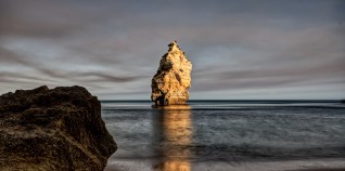 Ein Strand, dessen Meer sich in der Ferne auflöst. Im Vordergrund befindet sich ein Felsen. Im Hintergrund befindet sich ein zweiter Felsen, auf dem sich das Licht des Sonnenuntergangs spiegelt.