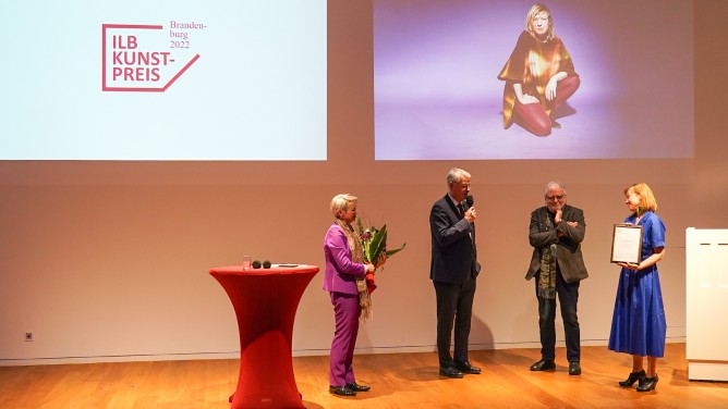 Es handelt sich um die Verleihung einer Auszeichnung. Es sind vier Personen. Eine hält einen Blumenstrauß, eine andere hält ein Mikrofon, während sie spricht. Eine andere Person hört aufmerksam zu und eine weitere Person zeigt eine Urkunde. Im Hintergrund sieht man das Porträt einer Künstlerin und das Logo des Preises, auf dem "ILB-Kunstpreis Brandenburg 2022" steht.