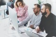 Bild eines Arbeitsplatzes, an dem ein Team bestehend aus einer Frau und zwei Männern, sich etwas auf einem Monitor anschauen.