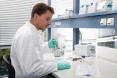 Mann sitzt im weißen Kittel am Labortisch mit Mikroskop und Pipette in der Hand. Daneben liege Unterlagen und Reagenzgläschen.