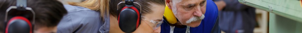 Maschinenwerkstatt in der eine junge Frau einem älterem Arbeiter etwas an einer Maschine zeigt, ein weitere, junger Mann schaut zu