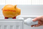 Orangefarbenes Sparschwein, auf welchem: "Heizkosten" steht, steht auf einem Konvektionsheizkörper. Eine Hand greift an das Thermostat.